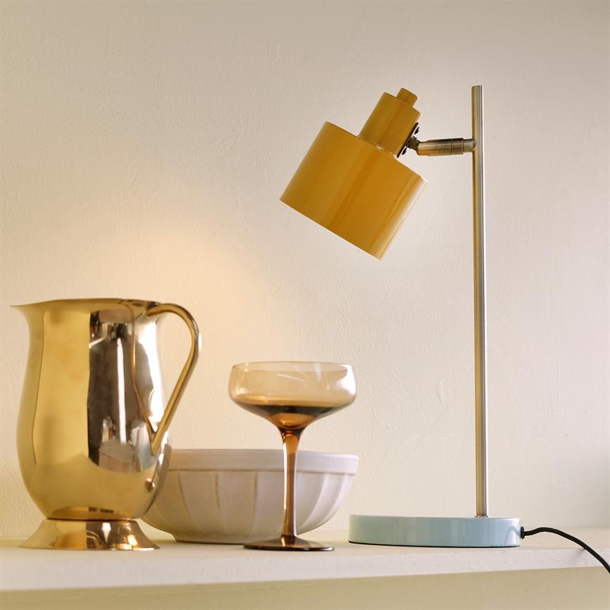 Gul bordlampe, vinklet nedad, kaster blødt varmt lys over en gylden kande, et ravfarvet cocktailglas og en hvid keramikskål på en lys hylde foran en cremefarvet væg.