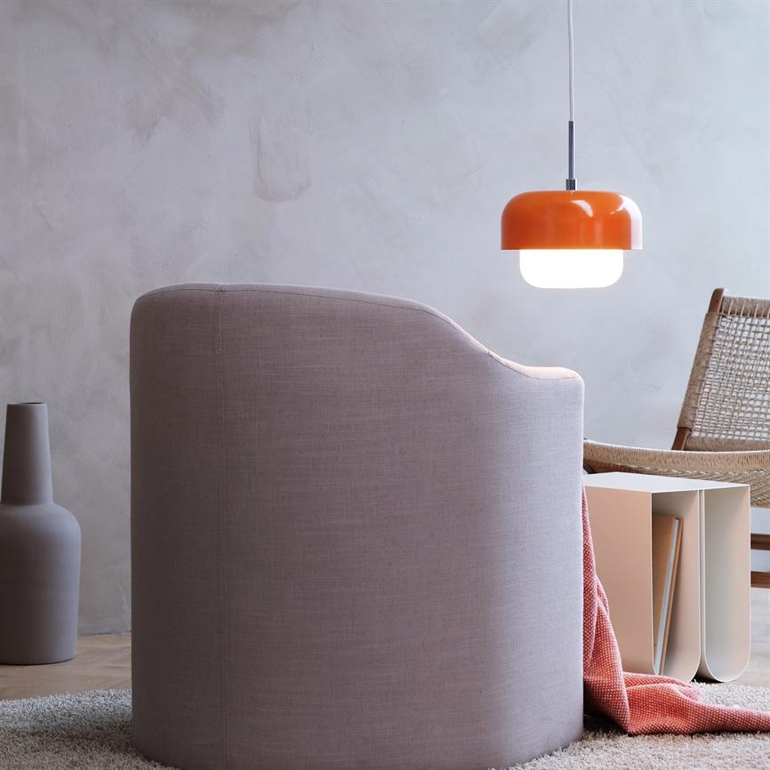En lyserød polstret lænestol vender væk, en orange hængelampe lyser over et lavt sidebord med bøger; en flettet stol, et tæppe og en vase står i et roligt, minimalistisk rum.