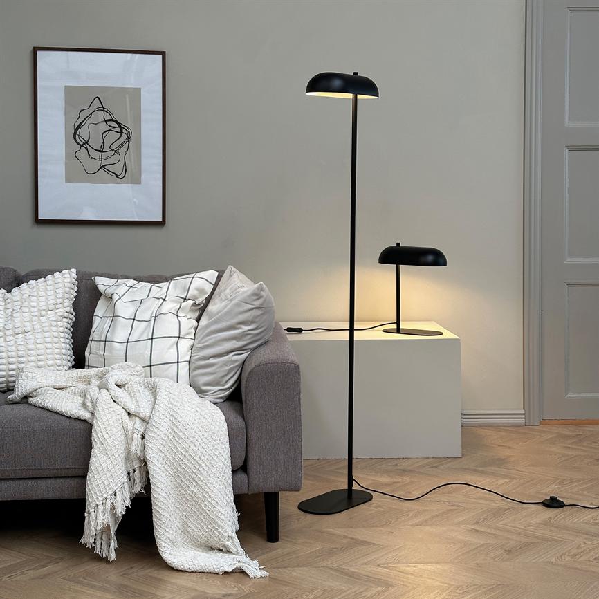 Gulvlampe står tændt og kaster varmt lys over en grå sofa med puder og et hvidt strikket tæppe, foran en hvid skænk med en mindre bordlampe og indrammet abstrakt kunst.