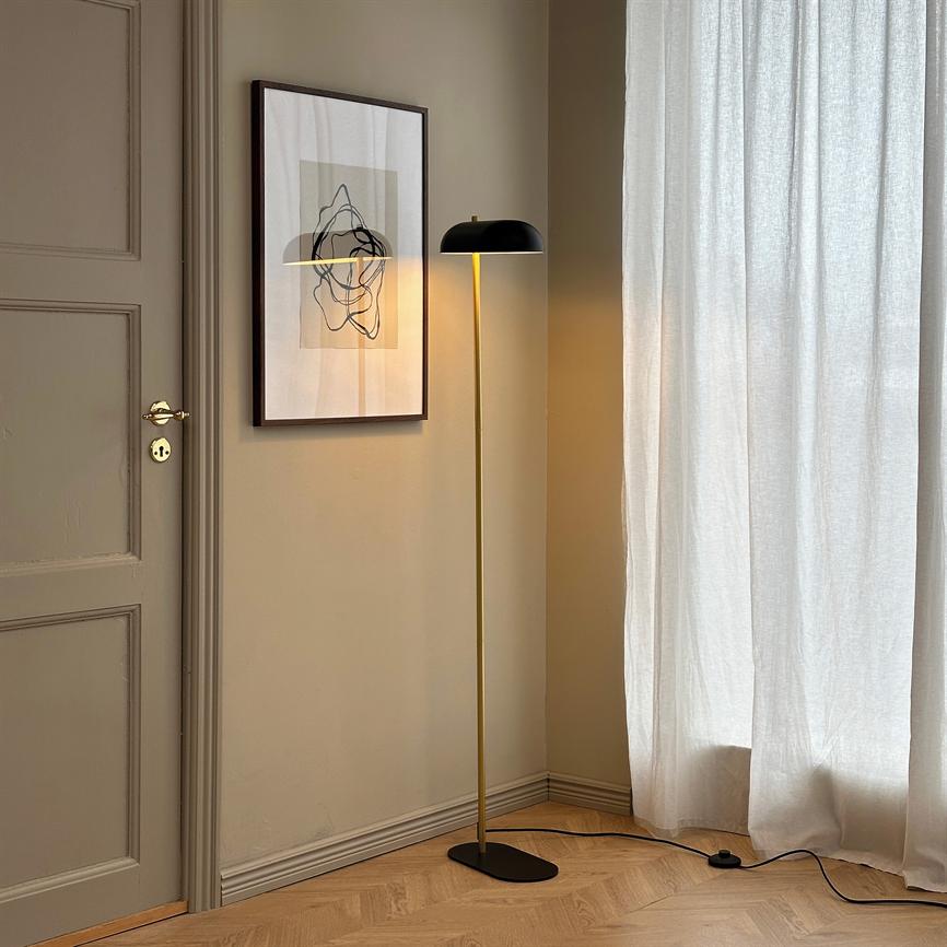 Gulvlampe står tændt og kaster varmt lys på et indrammet abstrakt billede; den står i et beige hjørne ved en lukket dør og et lyst gardin over parketgulv.