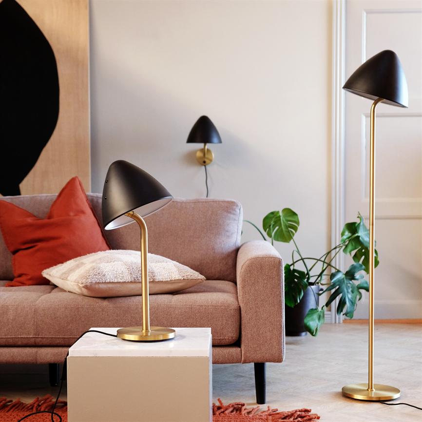 Bordlampe med sort skærm og messingstativ står på et hvidt sidebord ved en rosa sofa; en gulvlampe, en væglampe og en stor grøn stueplante ses i baggrunden.