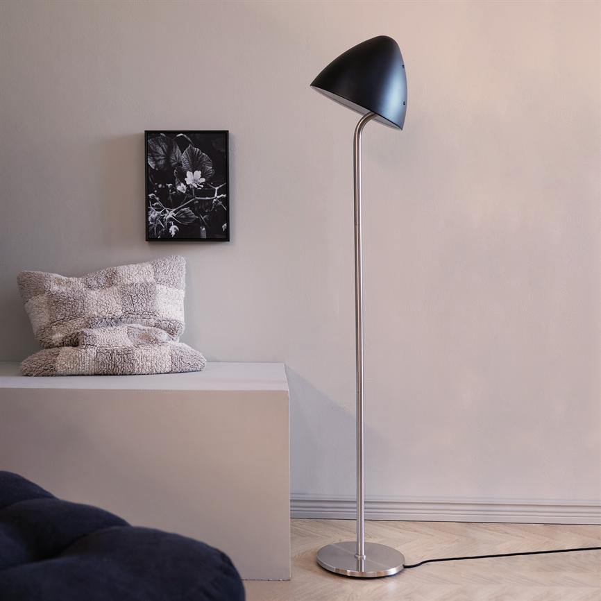 Gulvlampe står ved en lav bænk med to bløde, teksturerede puder, foran en beige væg med et lille sort-hvidt blomsterbillede; lyst trægulv og en sort ledning på gulvet.