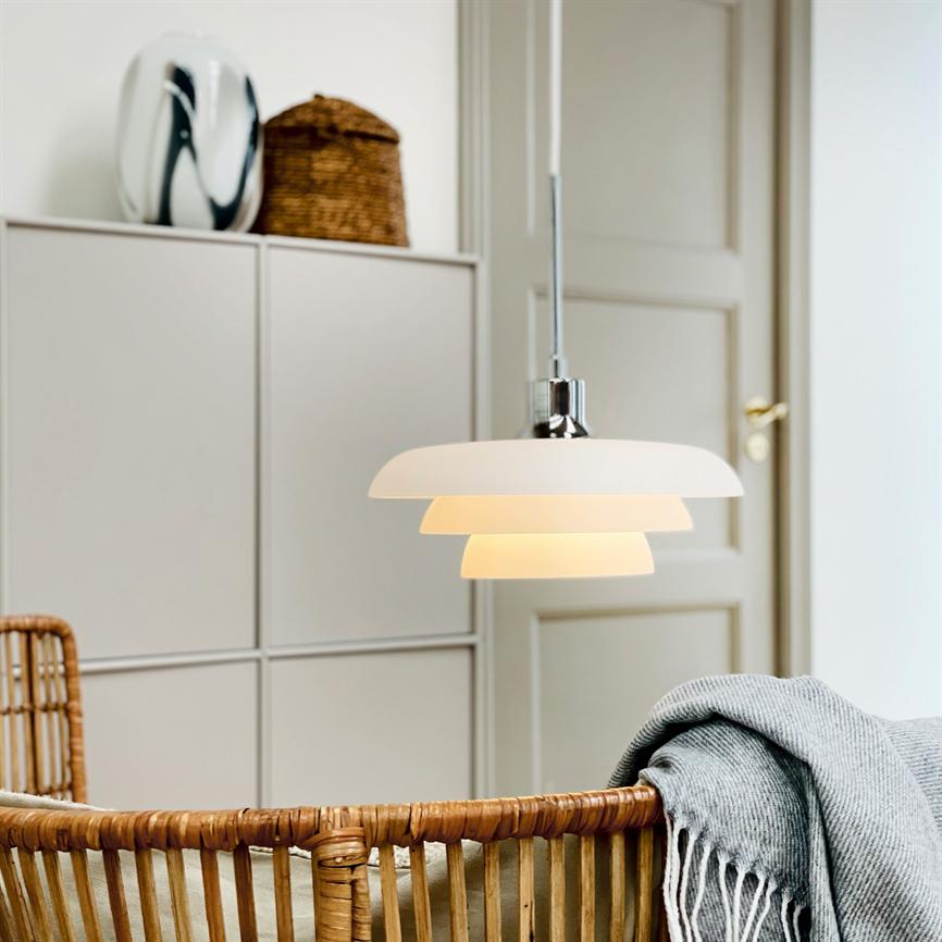 En hvid, lagdelt loftlampe hænger og udsender blødt, varmt lys over en fletstol i rattan med et gråt, frynset tæppe, foran neutrale skabe og en lukket dør i et lyst rum.