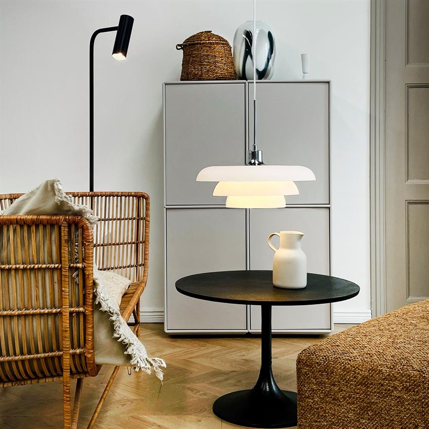 En hvid pendel lyser over et rundt sort bord med en hvid kande; i et lyst stuehjørne med flettet kurvestol, skab, stålampe og sildebensparketgulv.