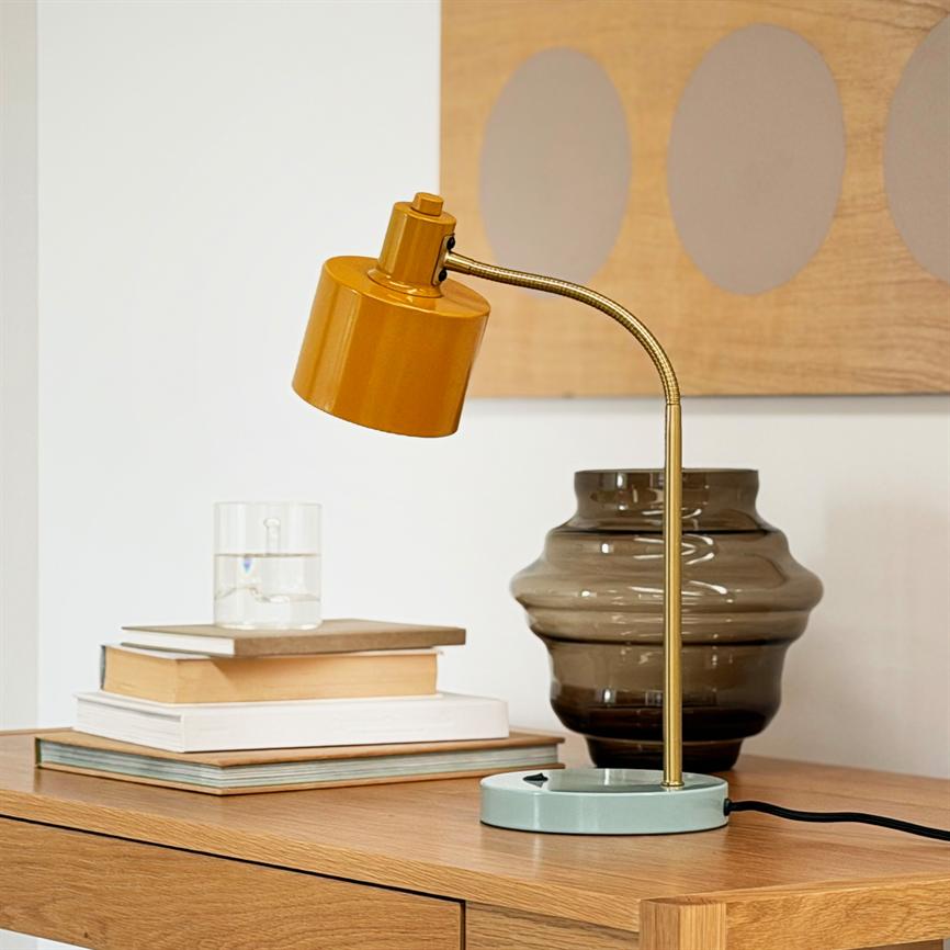 En okkerfarvet bordlampe med buet guldfarvet skaft står på et træbord ved en stak bøger og en røgfarvet glasvase i et lyst, minimalistisk rum med vægdekoration.