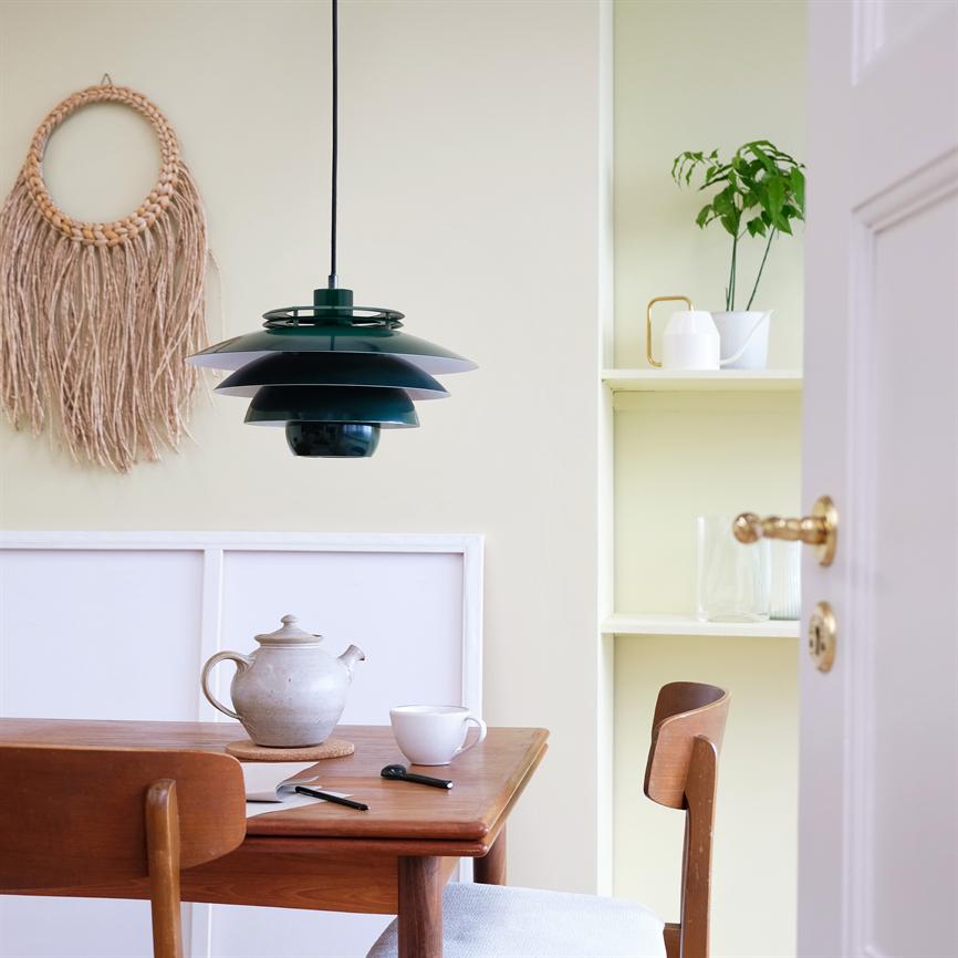En grøn skærmloftslampe hænger over et træbord med tekande, kop og notesbog; i en lys spisekrog med makramé-vægophæng, indbygget hylde med plante og delvist åben dør.