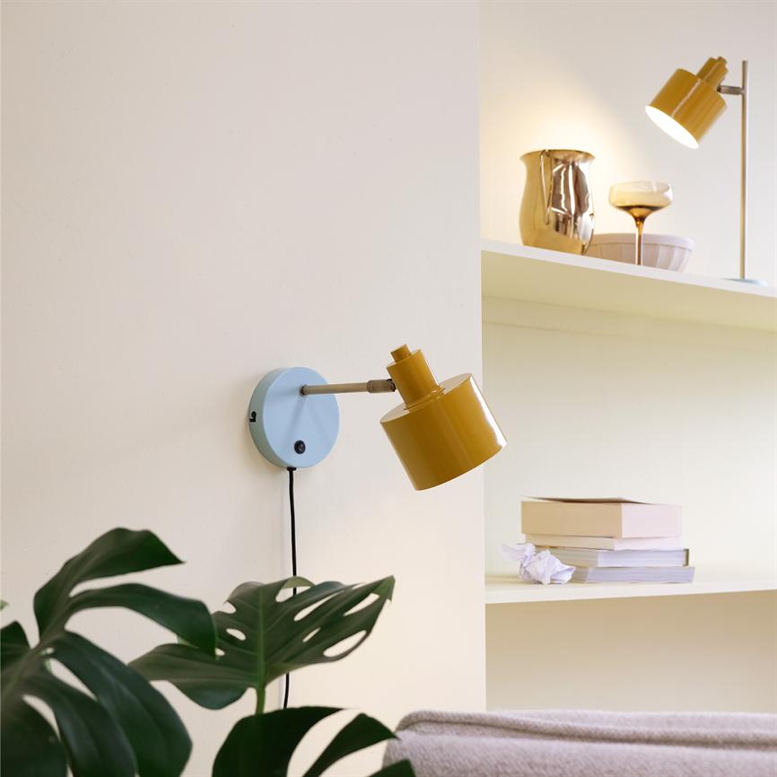 En sennepsgul væglampe monteret på en lyseblå plade peger nedad over en sofa, omgivet af en beige væg, en hylde med bøger og dekorative genstande og en grøn plante.
