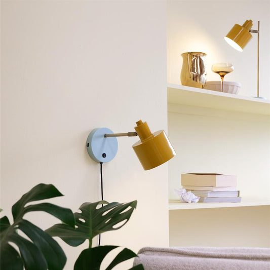 En sennepsgul væglampe monteret på en lyseblå plade peger nedad over en sofa, omgivet af en beige væg, en hylde med bøger og dekorative genstande og en grøn plante.