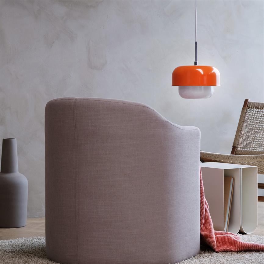 En lyserød polstret lænestol står vendt væk fra betragteren; et koralfarvet tæppe falder ned ved siden af. Minimalistisk stue med orange pendellampe, flettet stol, keramisk vase og sidebord.