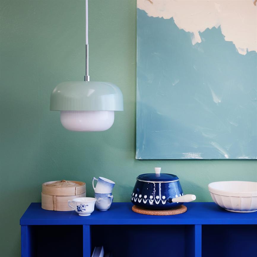 Lampe hænger over et blåt skab; en blå mønstret gryde står på skabet med kopper, en dampkurv og en hvid skål foran en grøn væg og abstrakt maleri.