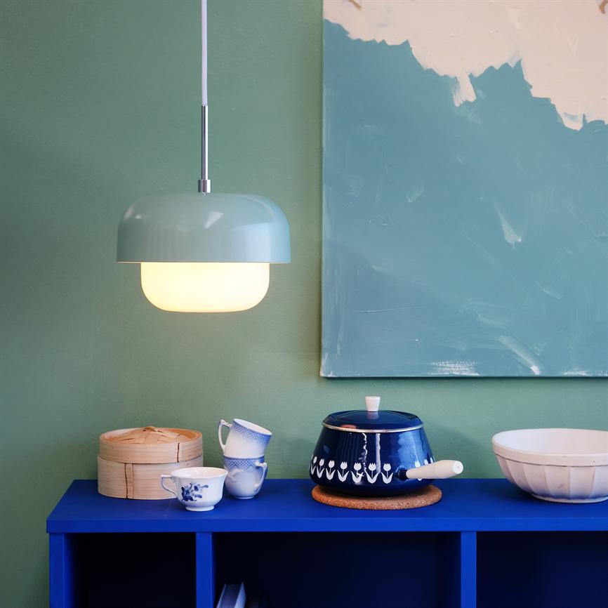 En ophængt pastelgrøn lampe lyser over en koboltblå kommode, hvor tekopper, bambusdampkurv og en mørk blå gryde står, foran en grøn væg med abstrakt maleri.
