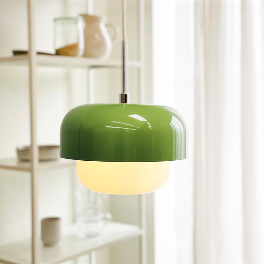 En grøn loftlampe hænger og udsender varmt lys i et lyst rum med hvide gardiner og en åben hylde med glas og keramik i baggrunden.