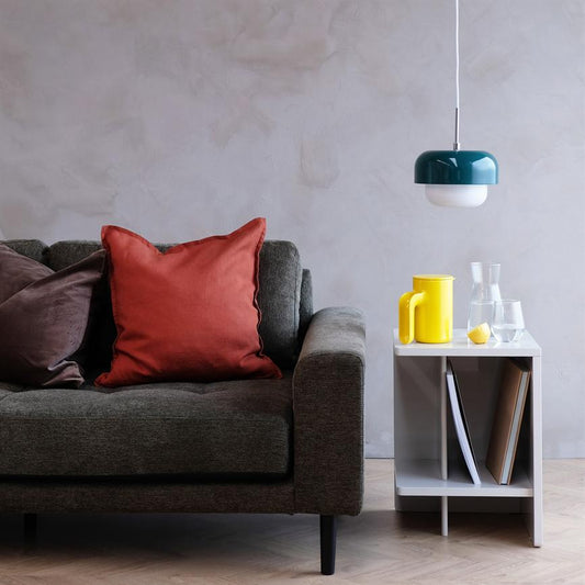 Grå sofa med en rød pyntepude, hviler roligt på lyst trægulv; ved siden står et hvidt sidebord med gul kande og glas, over det hænger en grøn loftslampe.