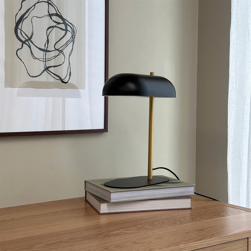 En sort og guld bordlampe står slukket på to stabelbøger på et træskab, foran en lys væg med indrammet abstrakt tegning og et hvidt, gennemsigtigt gardin.
