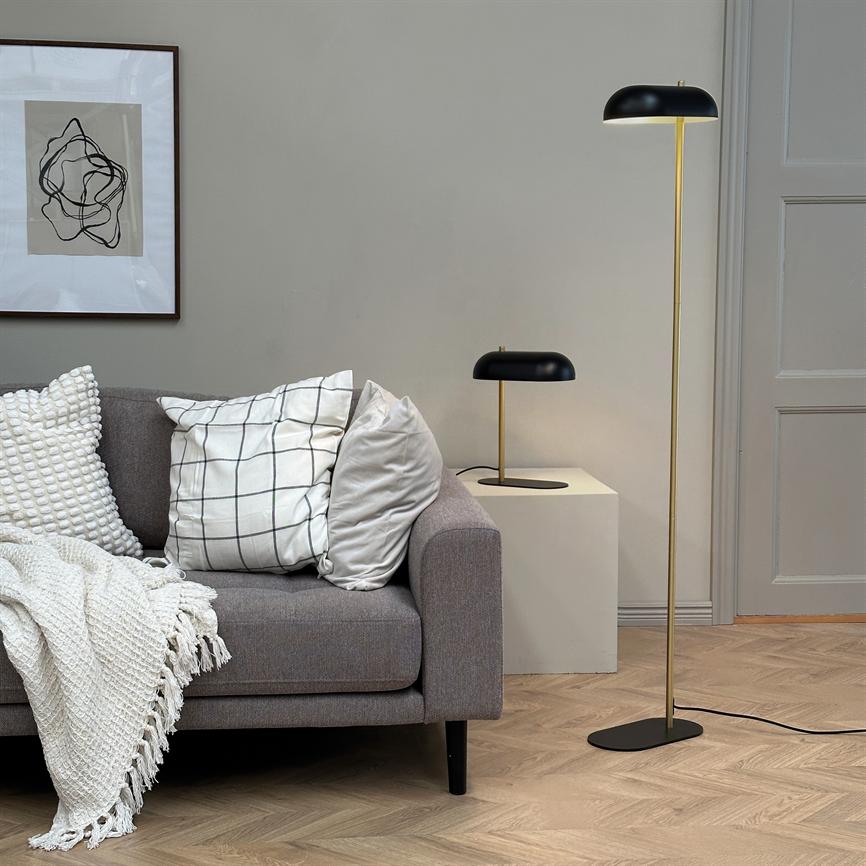 Grå sofa ligger med ternet og teksturerede puder og strikket tæppe; en sort-guld gulvlampe og bordlampe står ved siden af under indrammet abstrakt billede i et lyst, moderne stuerum.