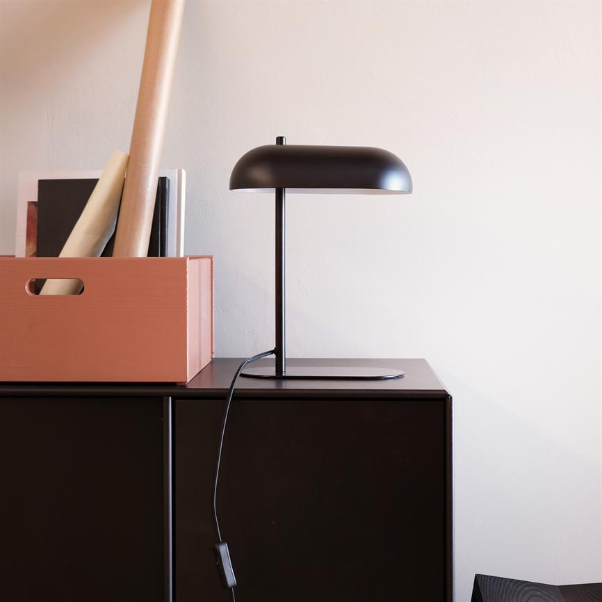 Sort bordlampe står på et mørkt skab, ledningen hænger ned; ved siden af en terrakotta opbevaringskasse med ruller og bøger, foran en lys, glat væg.