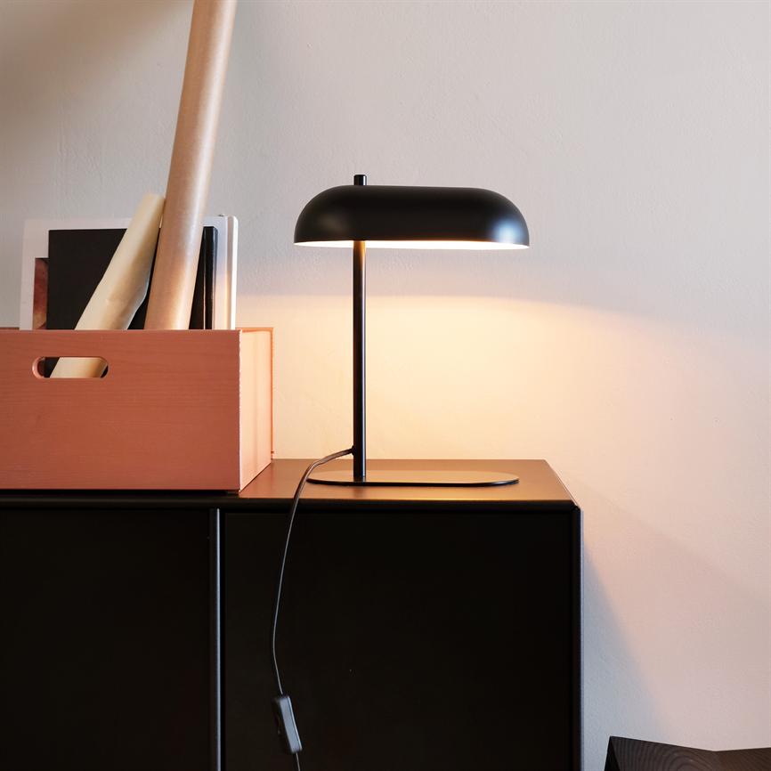 Sort bordlampe kaster varmt lys over et sort skab, står på en tynd fod; en sort ledning hænger ned langs skabets front; til venstre en orange opbevaringskasse med mapper og rullepapir.