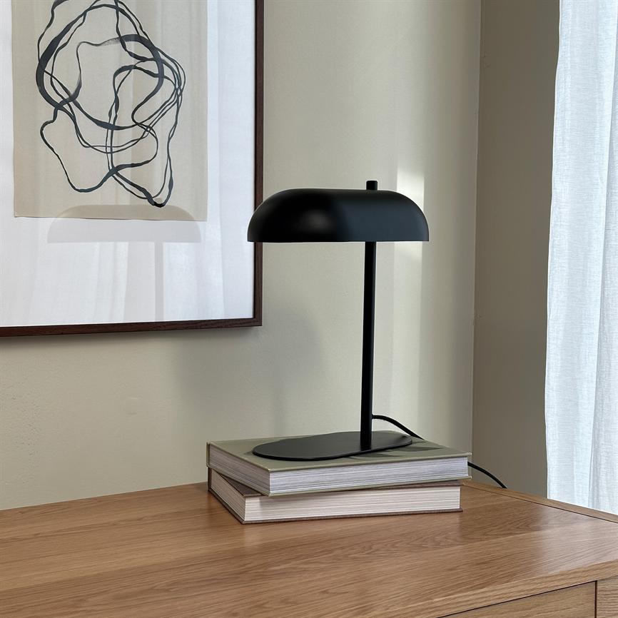 Sort bordlampe står på to stabler bøger på et træbord; blød naturlig belysning, neutral væg med indrammet abstrakt tegning til venstre og hvidt gardin til højre.