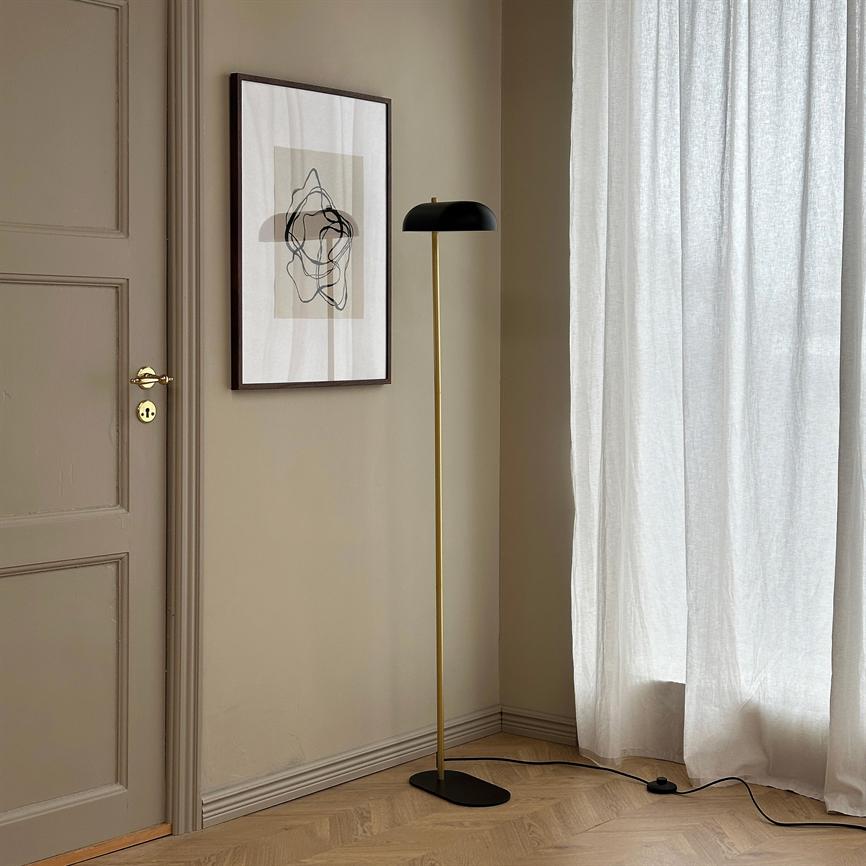 Gulvlampe med slank guldfarvet stang og sort kuppelskærm står i et hjørne; omkring den ses beige vægge, abstrakt indrammet plakat, en dør, lyse gardiner og parketgulv.