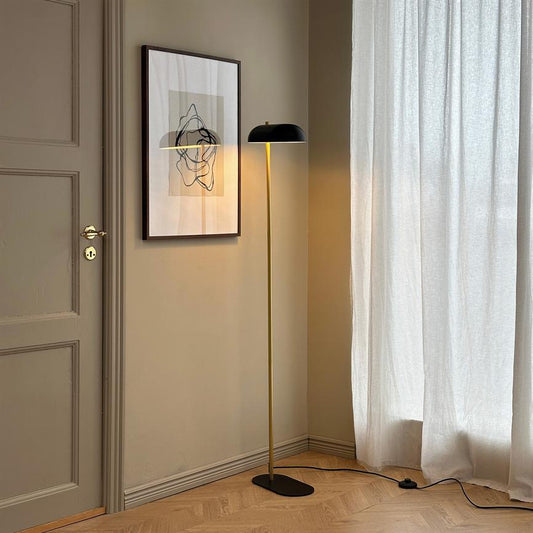 Gulvlampe står tændt og kaster varmt lys på et indrammet abstrakt billede; den står i et beige hjørne ved en lukket dør og et lyst gardin over parketgulv.