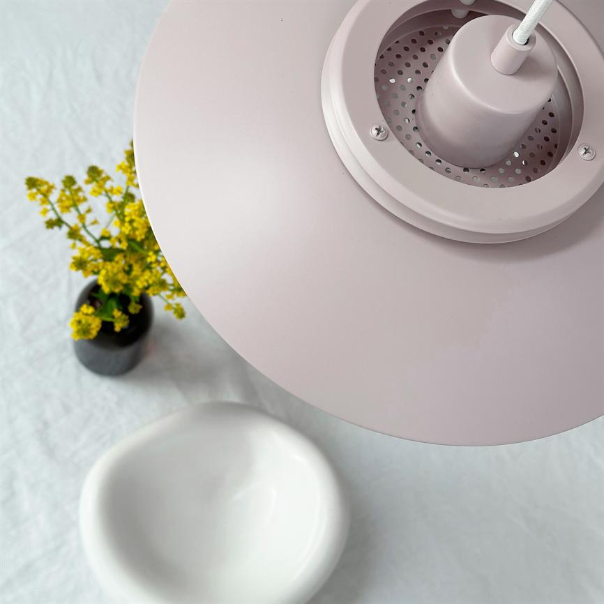En lyserød pendellampe dominerer forgrunden; den hænger over et bord med en lille vase med gule blomster og en hvid, oval skål på en lys dug.