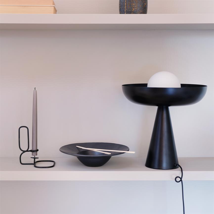 Sort konisk bordlampe med hvid glaskugle står til højre; midt på hylden hviler en sort skål med to lyse spisepinde; til venstre en grå stearinlys i sort metalstager. Hvid væg.