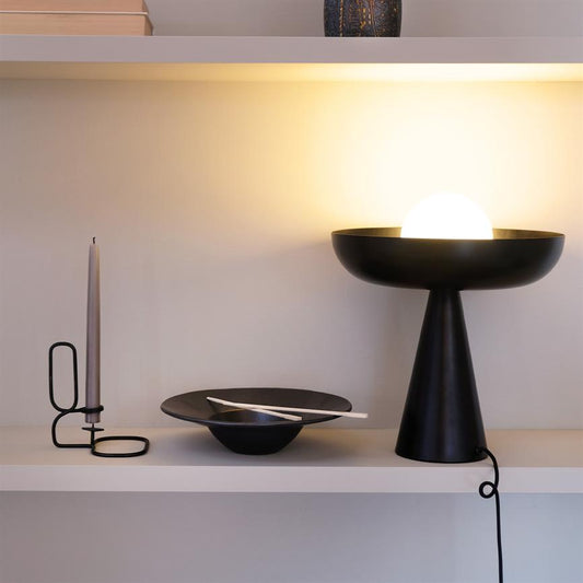 Sort konisk lampe udstråler varmt lys fra en kuglepære i en skål; står på en hvid hylde ved et slankt stearinlys i sort holder og en sort skål med hvide spisepinde.