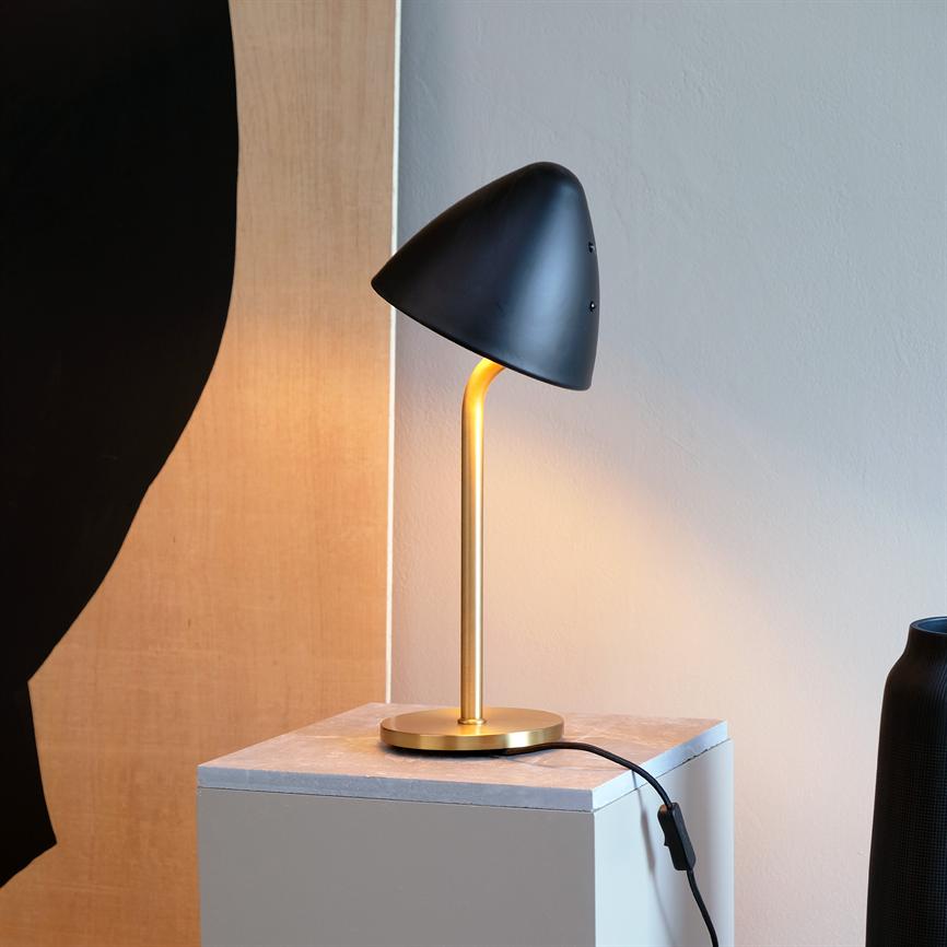 Sort, svampeformet bordlampe udsender varmt lys fra en gylden buet stander; den står på en lille firkantet sokkel foran en lys væg og træpanel, med en sort ledning synlig.