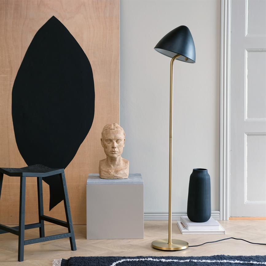 En gulvlampe med gylden stander og sort kuppel står ved en lertorso på en grå piedestal, flankeret af en sort vase, en skammel og et træpanel med sort abstrakt form.