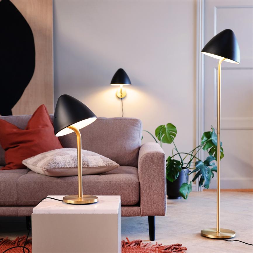 En sort og guldfarvet bordlampe lyser ved siden af en pudet sofa; suppleret af en matchende gulvlampe og væglampe, omgivet af planter, puder og et varmt, hyggeligt opholdsrum.