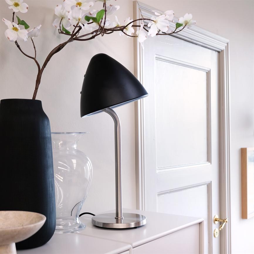 Bordlampe peger skråt nedad, placeret på et skab med metalfod og sort skærm, stående ved siden af en sort og en klar vase foran en hvid dør i et lyst rum.