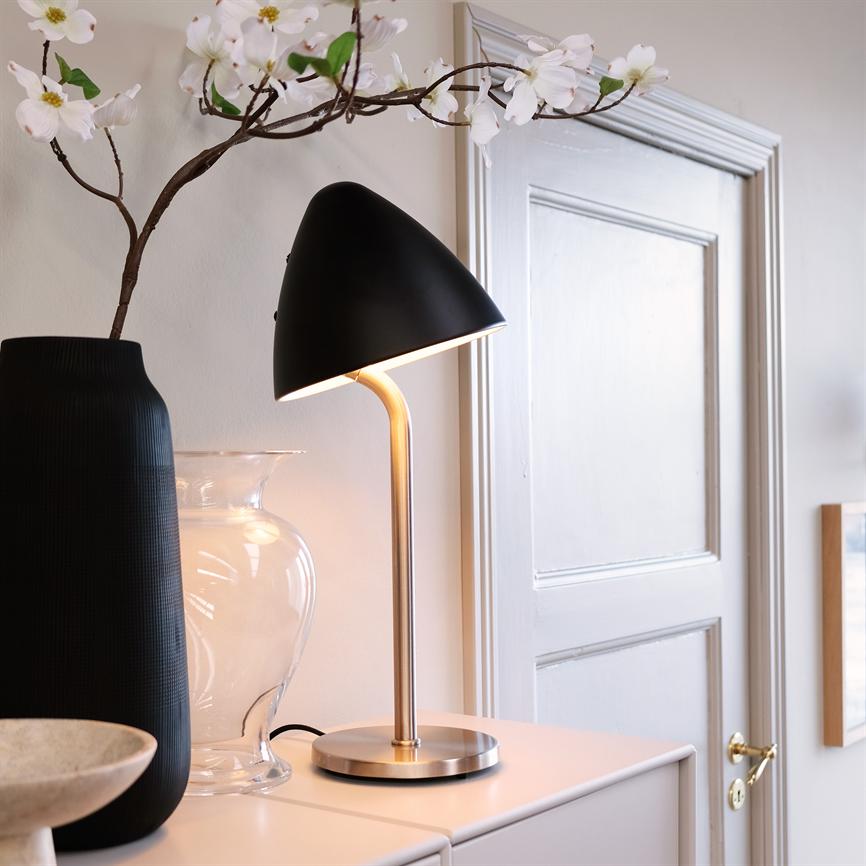 En sort bordlampe med messingstamme lyser blødt på et hvidt skab, flankeret af en høj sort vase, en klar glaskrukke og en blomstret gren foran en hvid dør.