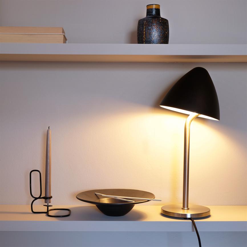 En sort bordlampe lyser varmt ned over en hvid hylde; ved siden af står en tynd lysestage, en sort skål med bestik, og en lille dekorativ vase på hylden ovenover.