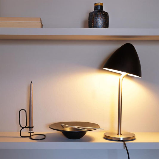 En sort bordlampe lyser varmt ned over en hvid hylde; ved siden af står en tynd lysestage, en sort skål med bestik, og en lille dekorativ vase på hylden ovenover.