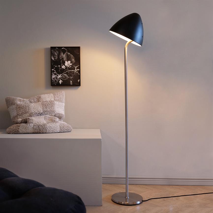 En slank gulvlampe lyser blødt mod en lys væg ved et lavt hvidt bord med en blød pude og et sort-hvidt plantebillede; parketgulv og synlig ledning.