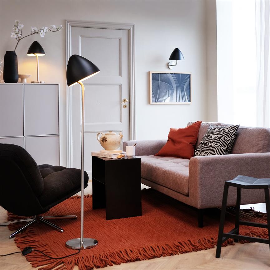 Gulvlampe står tændt ved en grå sofa; en sort lænestol, sort sidebord med keramikkande og puder på rødt tæppe i et lyst, minimalistisk stuehjørne.