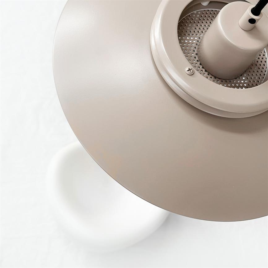 En beige metalpendel svæver over en rund hvid stol; lampeskærmen vises tæt på, monteret på sort ledning, i et lyst, minimalistisk rum.