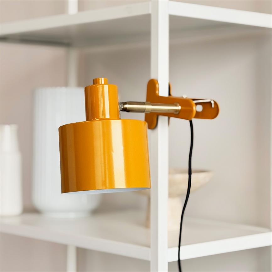 Orange klemelampe sidder fast på en hvid reol, hænger let skråt med sort ledning nedad, lyser ikke; baggrunden viser udviskede hvide vaser og neutrale hylder.