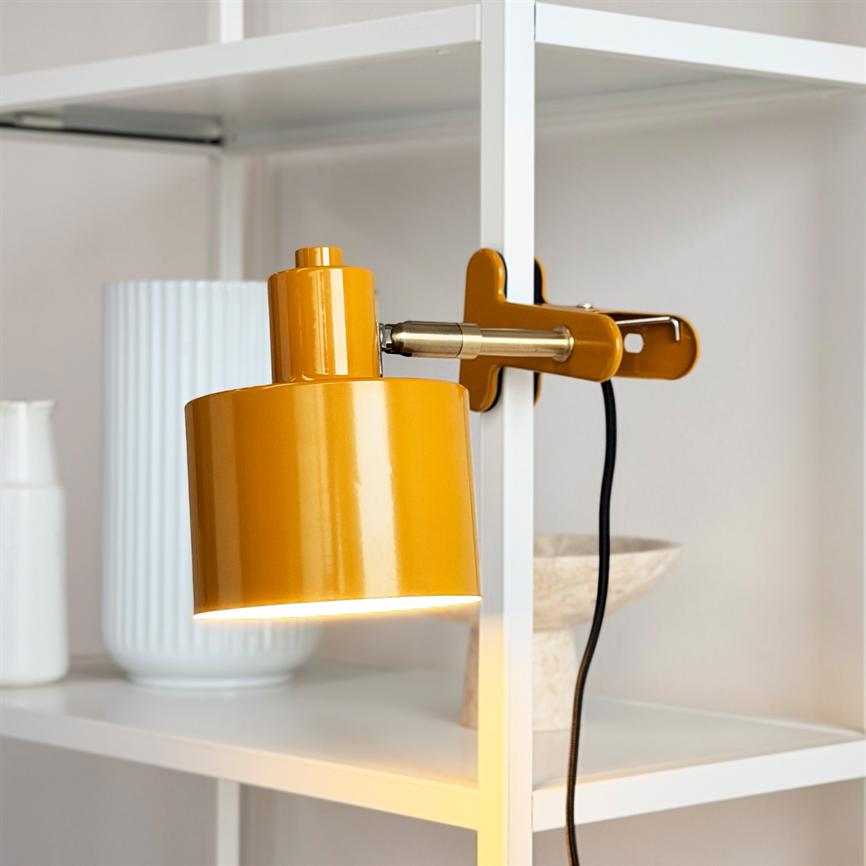 Gul klemme-lampe er tændt og lyser nedad, monteret på en hvid reol med hvide keramikvaser og en skål i baggrunden; sort ledning hænger ned.
